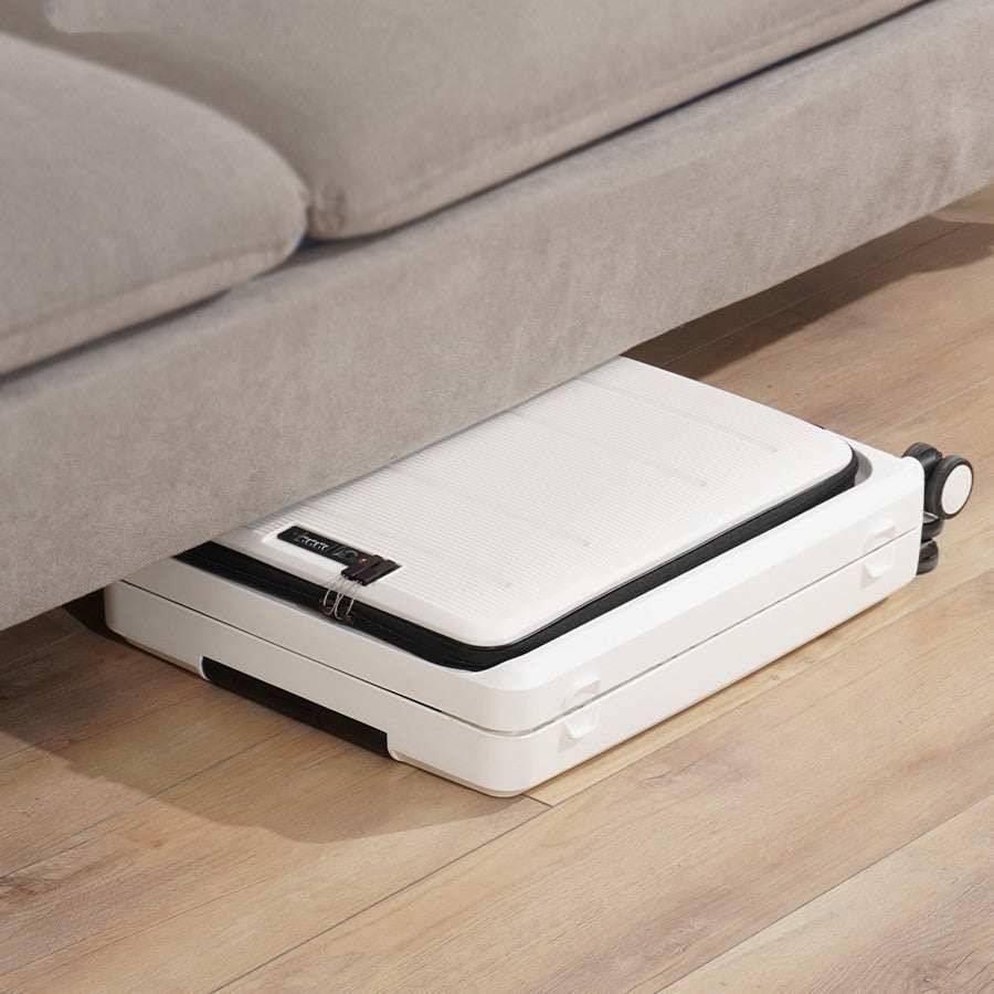 Foldable white luggage stored under sofa with zipper closure and aircraft wheels.