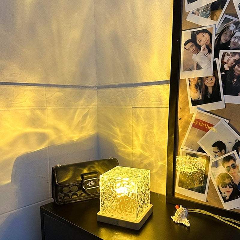 LED Water Ripple Ambient Night Light projecting warm yellow water ripple effect on wall with crystal cube design on black table next to mirror and photo collage.